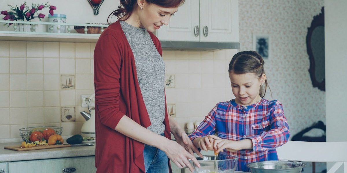 Little cheerful girl helping her mother in the kitchen cropping egg into bowl and mixing dough for cookies. Family, food, home and people concept