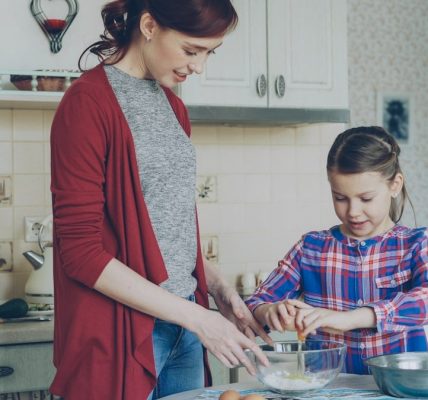 Little cheerful girl helping her mother in the kitchen cropping egg into bowl and mixing dough for cookies. Family, food, home and people concept