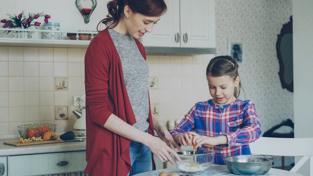 Little cheerful girl helping her mother in the kitchen cropping egg into bowl and mixing dough for cookies. Family, food, home and people concept